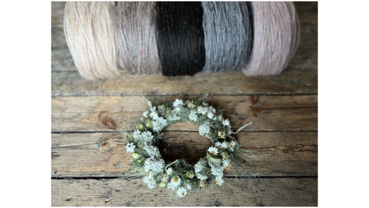 A rustic wooden floor serves as a backdrop for a beautifully crafted flower crown, with an assortment of colorful plates of unspun Nutiden yarn from Höner och Eir arranged in the soft-focused background.