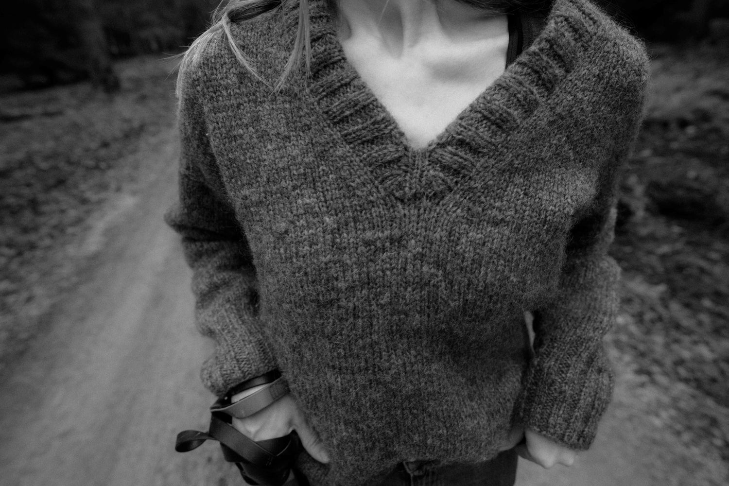 A black and white close-up photo of a person wearing a cozy, hand-knitted V-neck sweater design 'Chloe Sweater'. The sweater, knit in Nutiden unspun yarn, is a modified drop shoulder design with long sleeves.