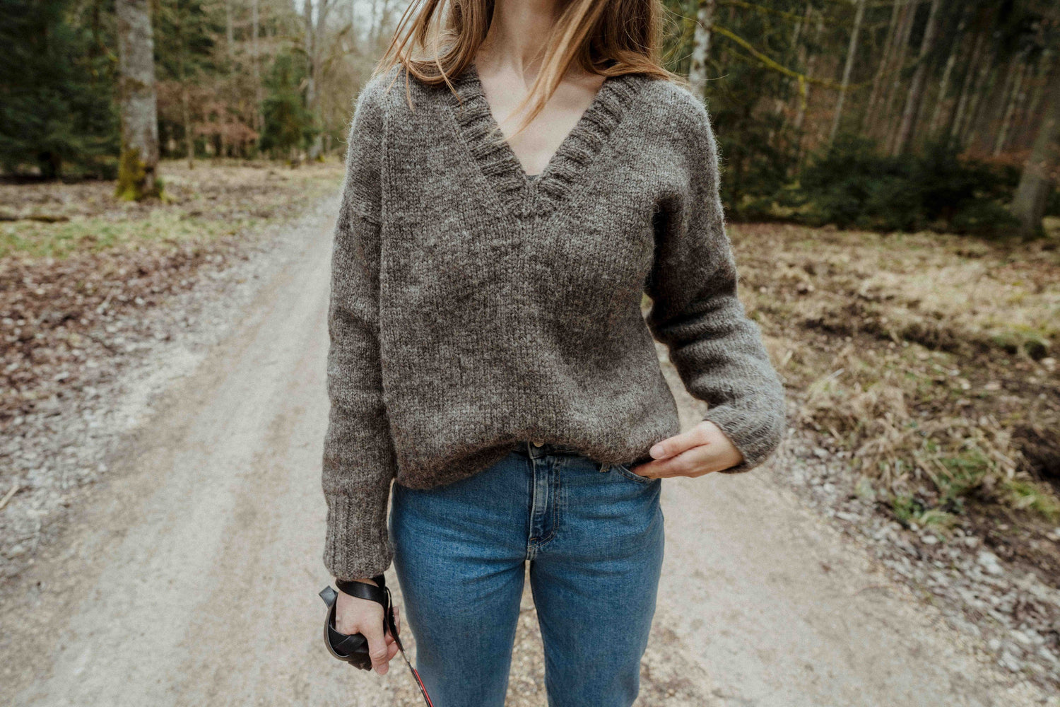 A close-up image featuring the designer Ulven, outdoors. She is wearing a hand-knitted V-neck sweater in a natural shade of medium grey paired with blue jeans. In her hand, she holds a dog leash, subtly indicating the presence of a pet out of frame. The background showcases a tranquil, forested path.