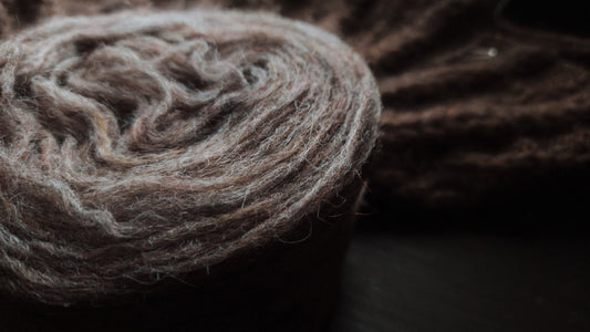 A single plate of pink brown unspun Nutiden yarn in the foreground on a dark brown table. In the background, a blurred image showcases the work-in-progress of a handknit Soft Spoken Sweater.