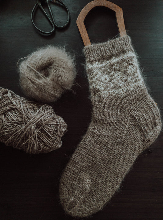 A beige knitted sock with colourwork pattern on a sock blocker, surrounded by yarn and scissors.