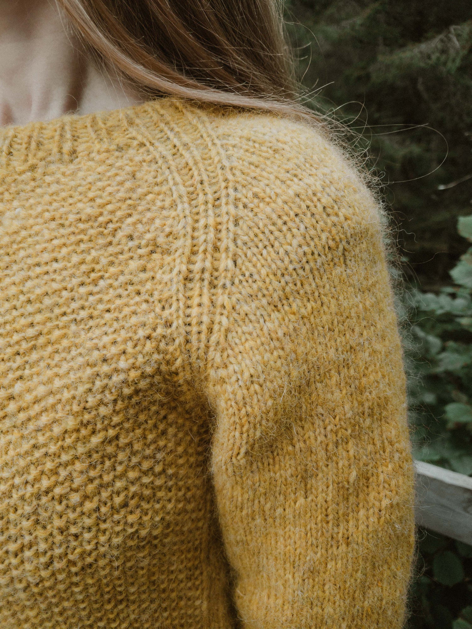 Close-up of the ribbed raglan seam and textured seed stitch on the 'Before Fall Sweater' by Ulven, presented in amber-yellow. The garment, worn by the designer, is set against a soft-focus background of green foliage and is crafted with unspun Nutiden yarn from Höner och Eir.