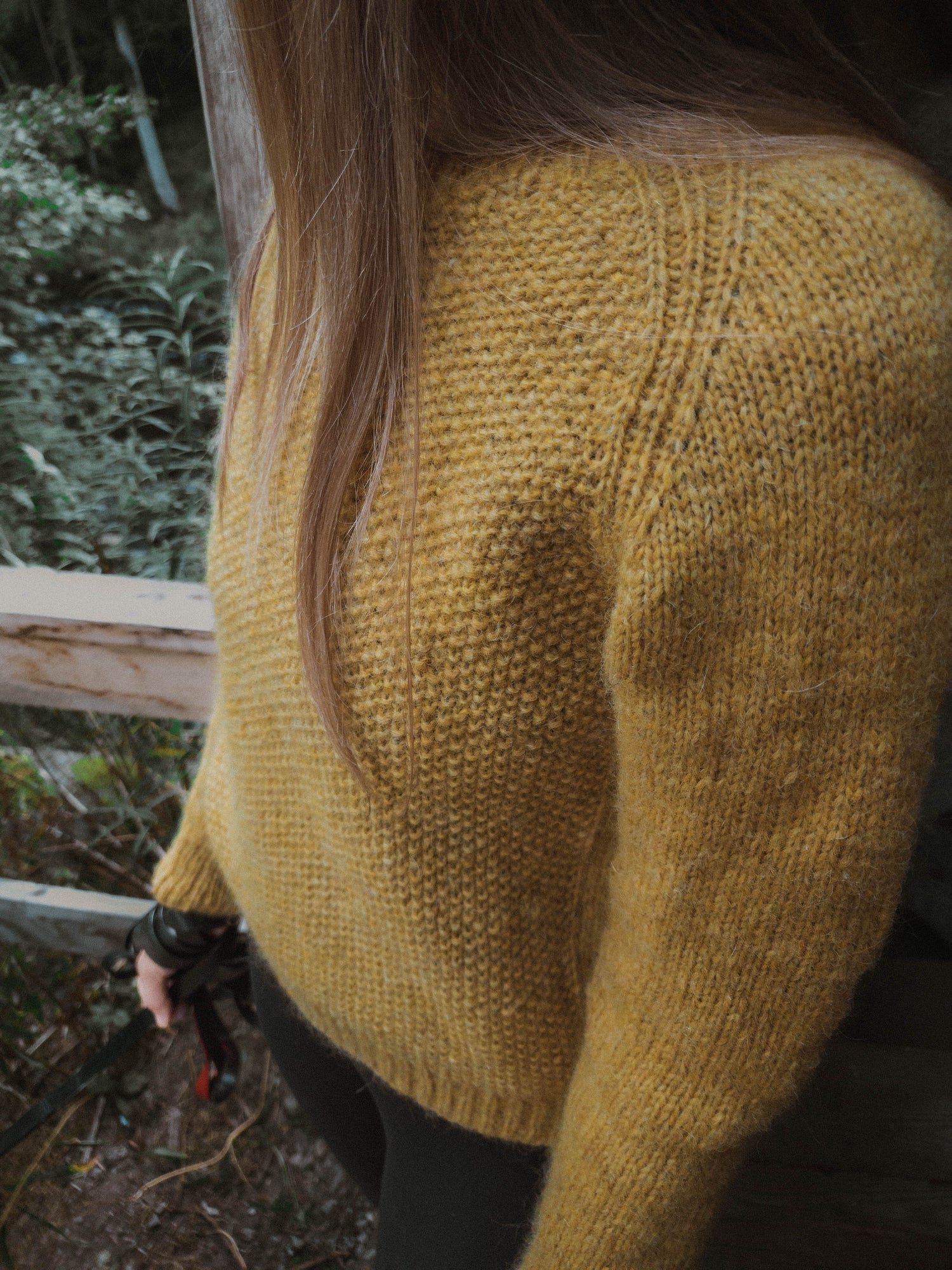 Close-up of the ribbed raglan seam and textured seed stitch on the 'Before Fall Sweater' by Ulven, presented in amber-yellow. The garment, worn by the designer, is set against a soft-focus background of green foliage crafted with unspun Nutiden yarn.