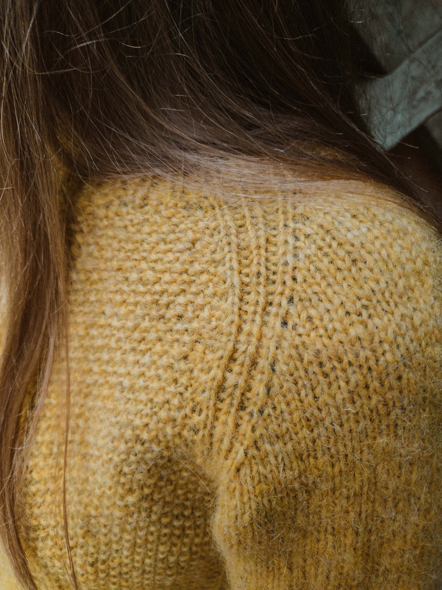 Close-up of the ribbed raglan seam and textured seed stitch on the 'Before Fall Sweater' by Ulven, presented in amber-yellow. The garment, worn by the designer, is set against a background of a wooden bridge, crafted with unspun Nutiden yarn.