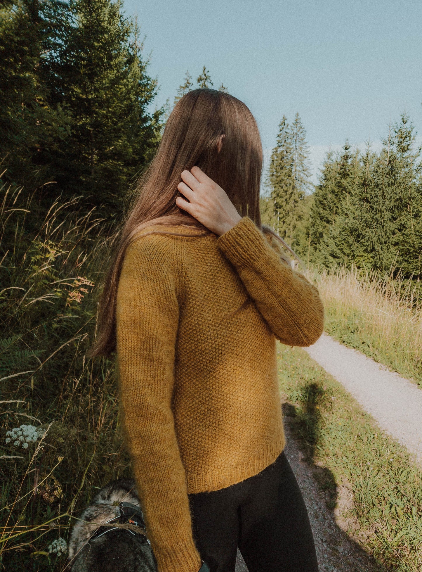 Full view of the 'Before Fall Sweater' by Ulven, worn by the designer against the serene backdrop of a forest road encased by trees on a late summer day. The amber-yellow sweater, knit with unspun Nutiden yarn from Höner och Eir, features a ribbed raglan seam and textured seed stitch, fully displayed in natural light.