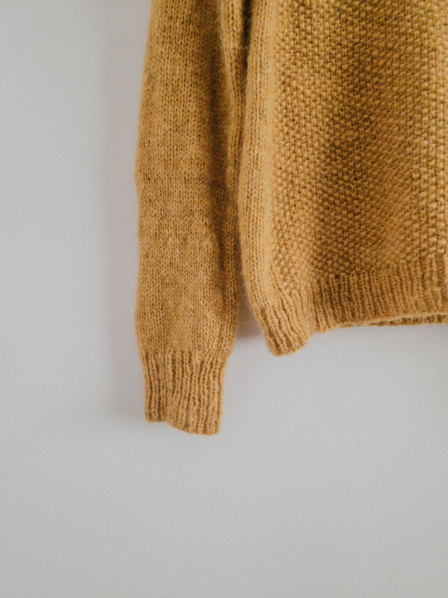 Close-up of the lower sleeve and hem of the hand-knit 'Before Fall Sweater' by Ulven, amber-yellow, showcased on a black hanger against a white backdrop. The intricate details of the unspun Nutiden yarn and the textured seed stitch pattern are highlighted.