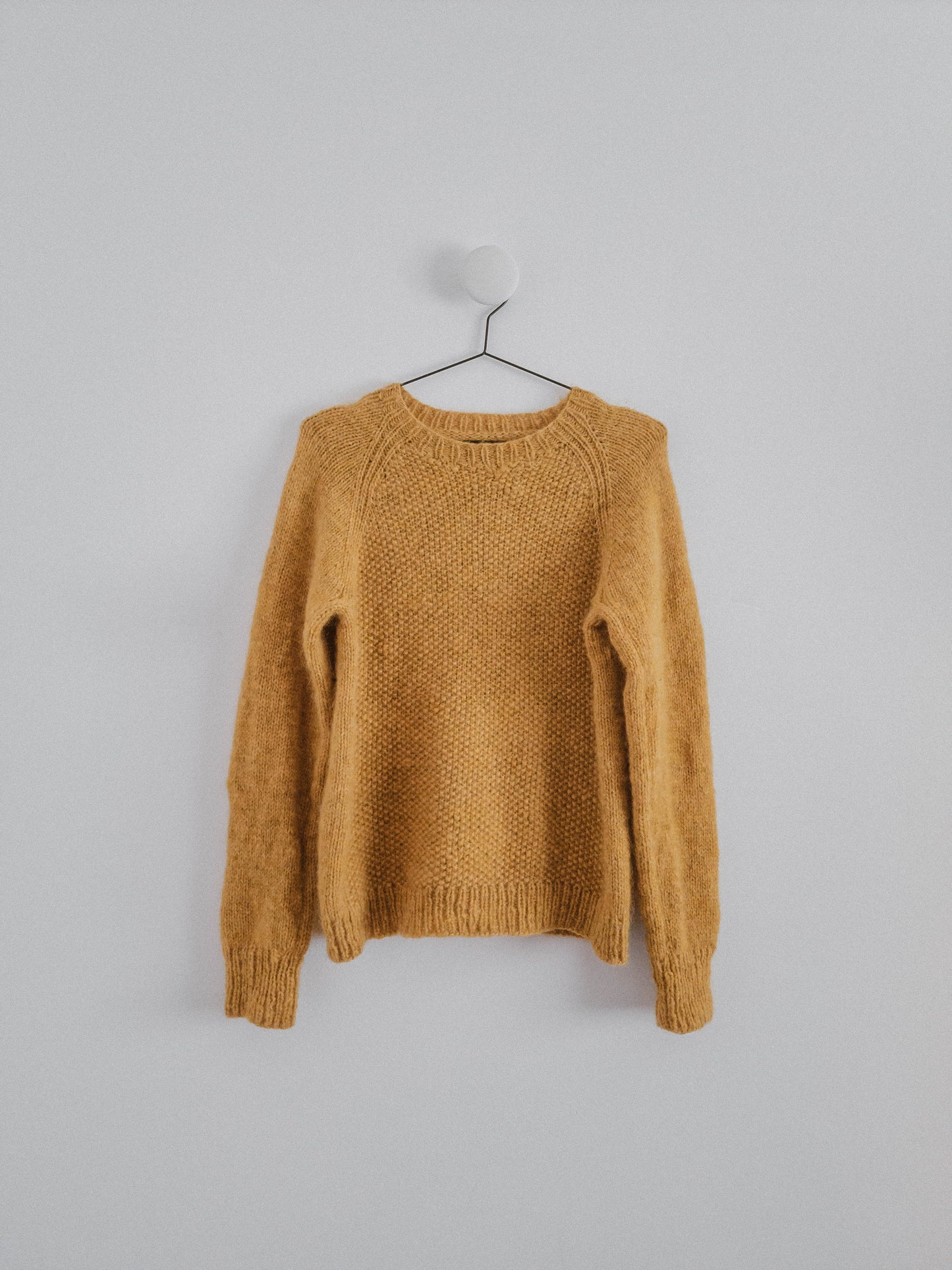 Amber-yellow hand-knit raglan 'Before Fall Sweater' by Ulven on a black hanger, white background. Made with unspun Nutiden yarn by Höner och Eir, featuring a seed stitch front panel.