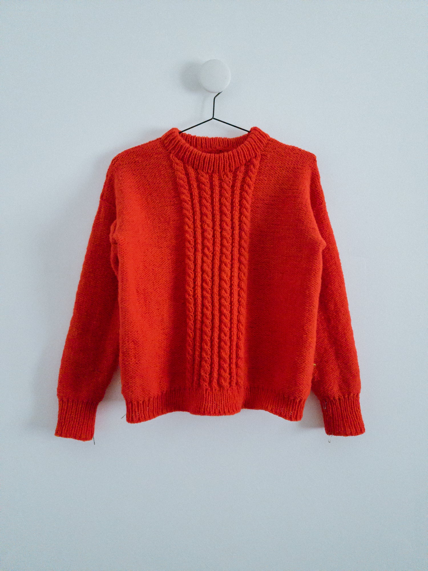 The HELIOS SWEATER designed by Lív Ulven, knitted in vibrant red Mota Wooldreamers yarn, hanging on a minimalist white wall with a simple hanger.