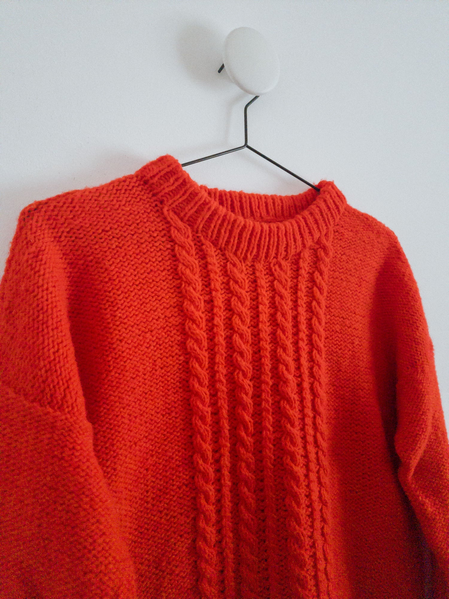 The HELIOS SWEATER designed by Lív Ulven, knitted in vibrant red Mota Wooldreamers yarn, hanging on a minimalist white wall with a simple hanger.