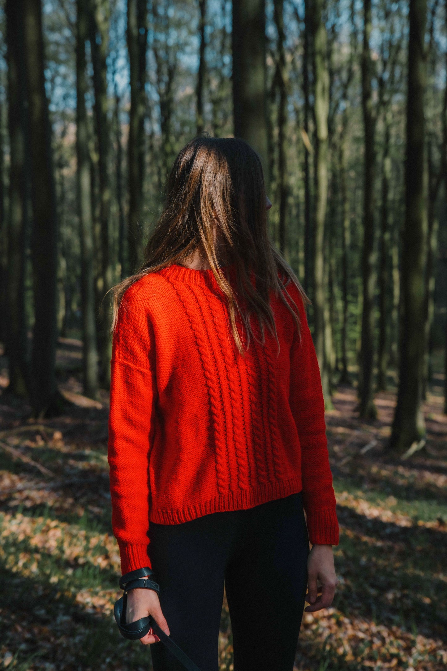 The designer wearing the HELIOS SWEATER, designed by Lív Ulven, knitted in vibrant red Mota Wooldreamers yarn, standing in a forest setting.