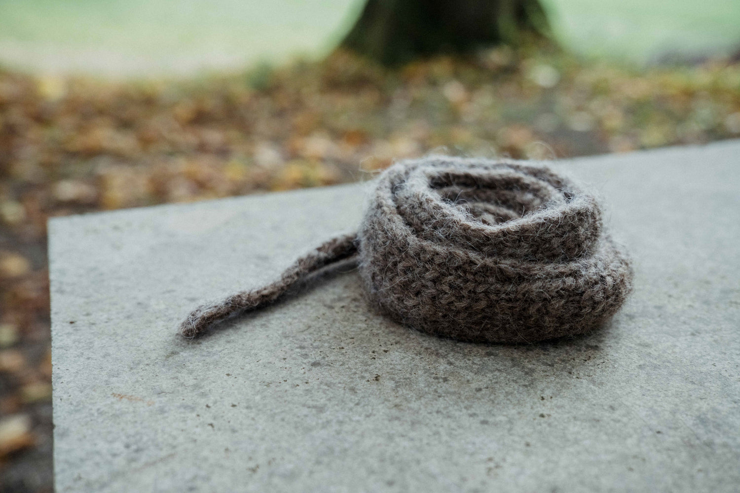 Grey-brown HAZEL SCARF with I-cord edge and slip-stitch, on concrete, Nutiden yarn, blurred autumn backdrop.