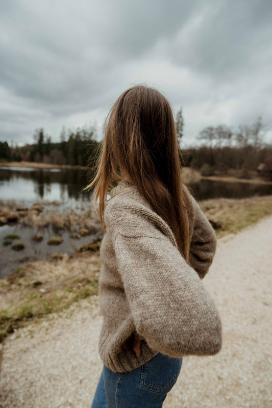 A serene, outdoor portrait featuring the designer, Ulven, from behind, looking out over a scenic lake. She is wearing a handmade, cozy beige sweater and blue jeans and holding a dog leash.