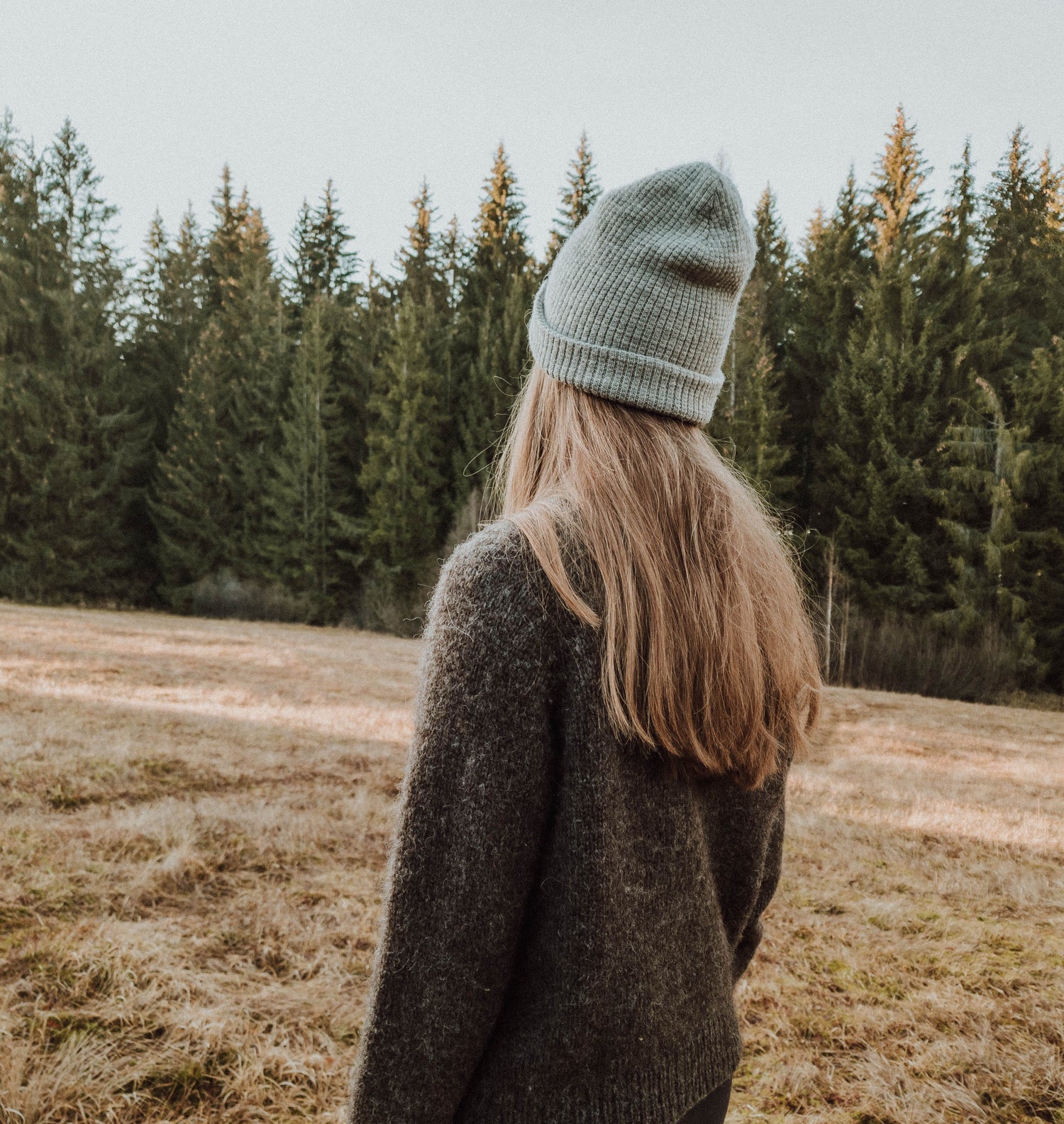 Knitwear designer Woodlandsknits long dark blonde hair, donned in a nature black hand-knitted sweater made from unspun yarn and a grey hat, standing in an alpine field, during sundown gazing towards the horizon graced by a line of tall trees.