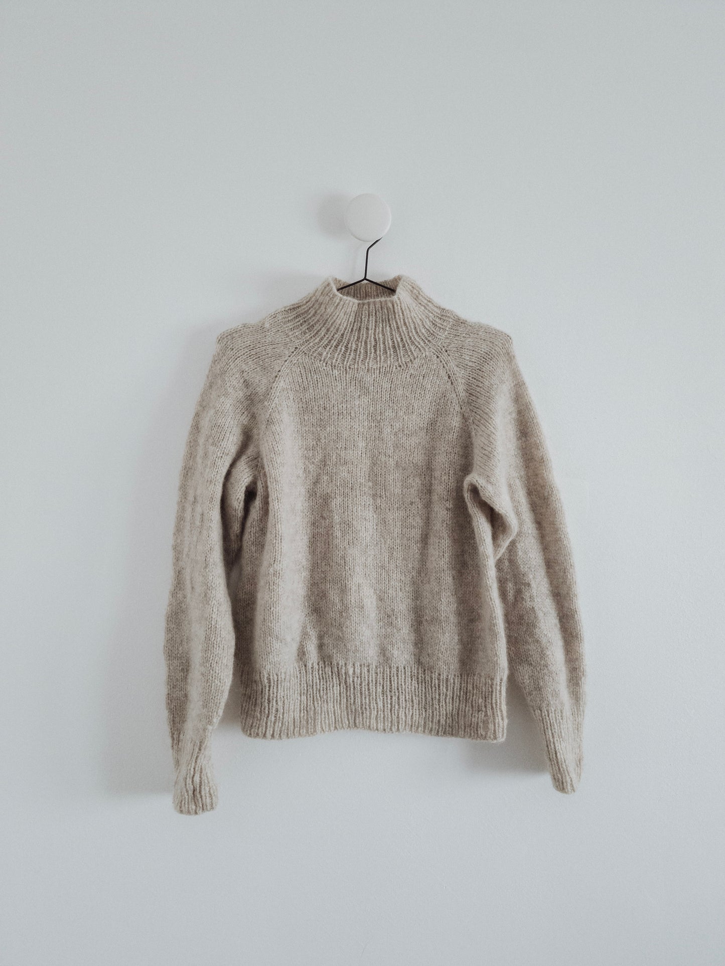 Minimalist photo of a very light grey, soft, hand-knit raglan sweater with a mock turtleneck, crafted by Woodlandsknits from Nutiden unspun yarn. The sweater is displayed on a black hanger against a white wall.